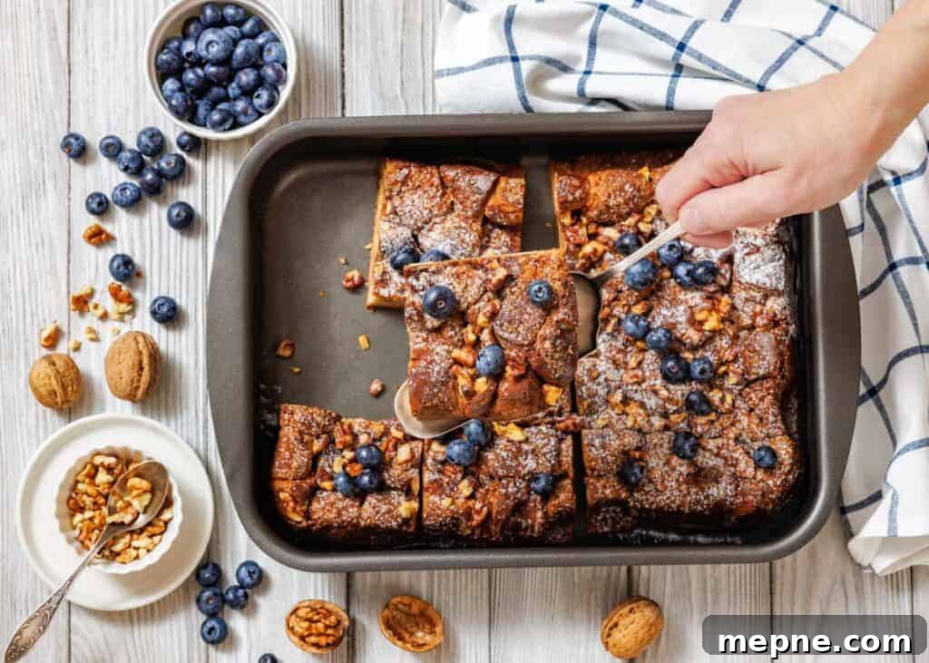 35 Classic Casserole Recipes That Have Everyone Coming Back 11 A baking pan with sliced Baked Cinnamon French Toast is shown, with a hand using a spoon to lift one piece.