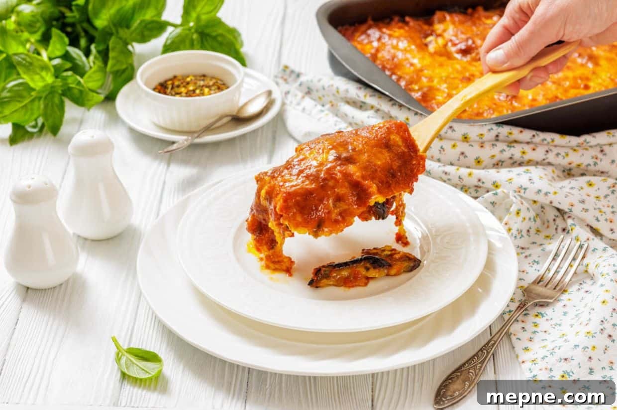 35 Classic Casserole Recipes That Have Everyone Coming Back 6 A hand serves a slice of Eggplant Parmigiana onto a white plate, with a casserole dish, herbs, and condiments visible on a white table.