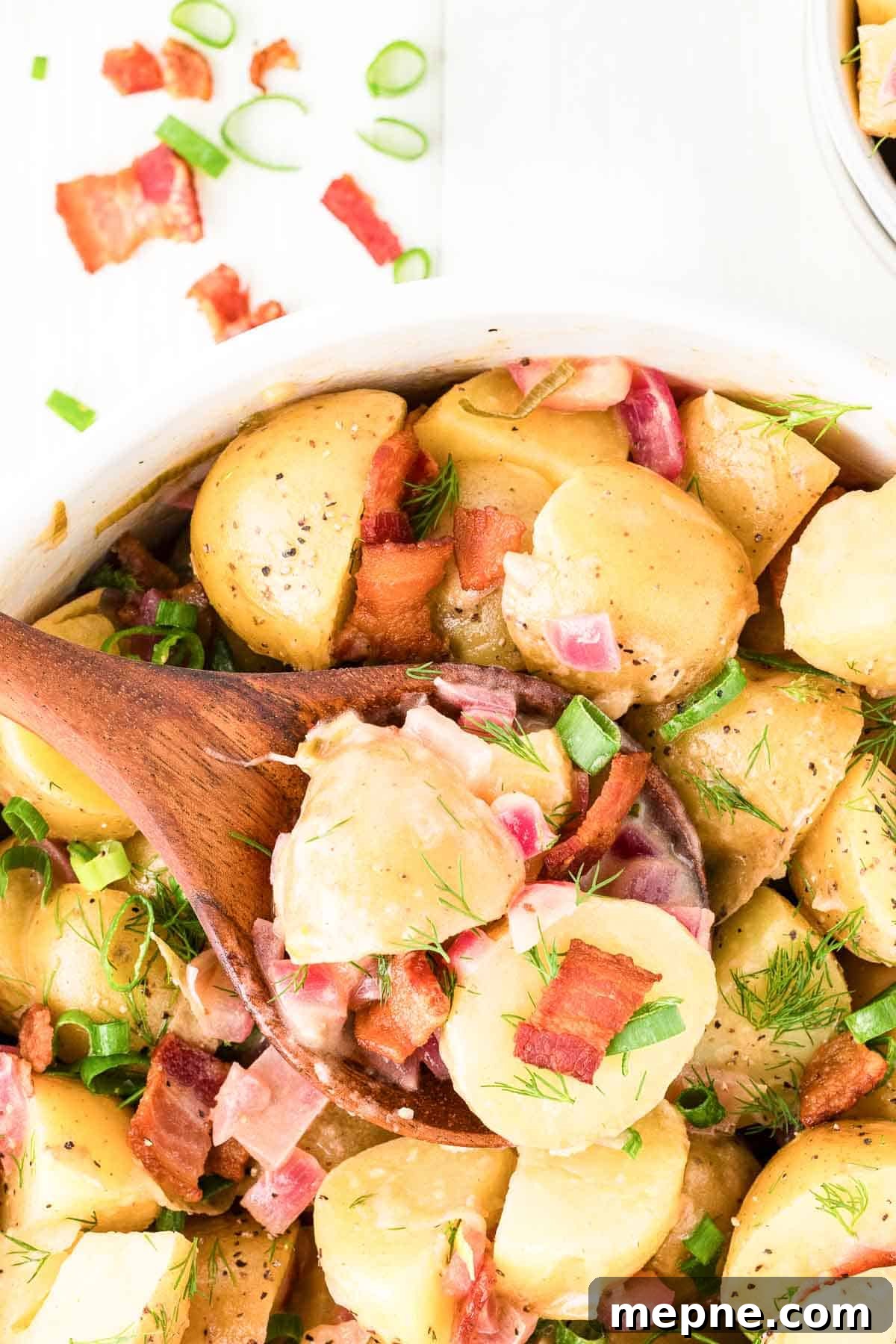 Authentic Bavarian Potato Salad with Bacon Recipe 2 A bowl of Bavarian potato salad with chunks of potatoes, bacon pieces, green onions, red onions, and dill, with a wooden spoon serving a portion.