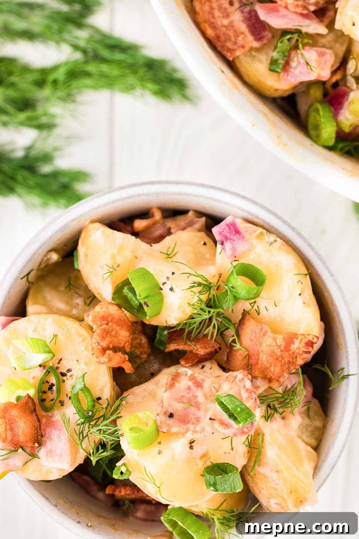 Authentic Bavarian Potato Salad with Bacon Recipe 19 A bowl of Bavarian potato salad garnished with dill, green onions, and bacon pieces is placed on a white wooden surface next to fresh dill.