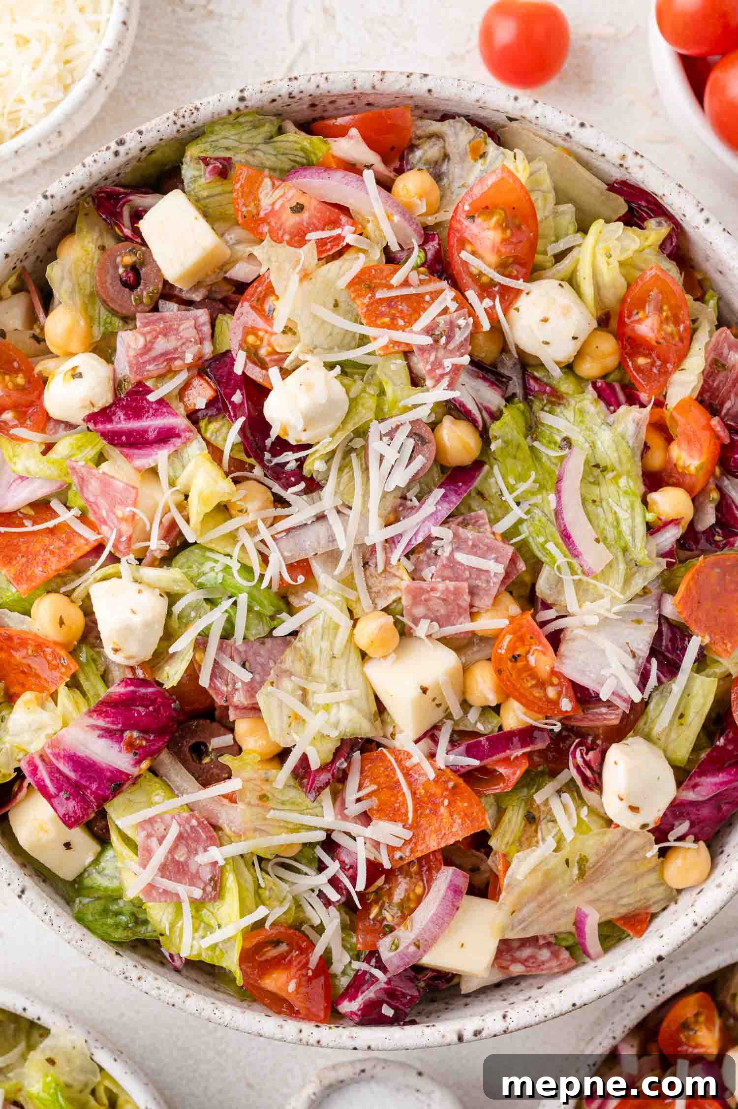 A bowl of chopped salad with lettuce, sliced salami, mozzarella cubes, cherry tomatoes, chickpeas, shredded cheese, and red onions.