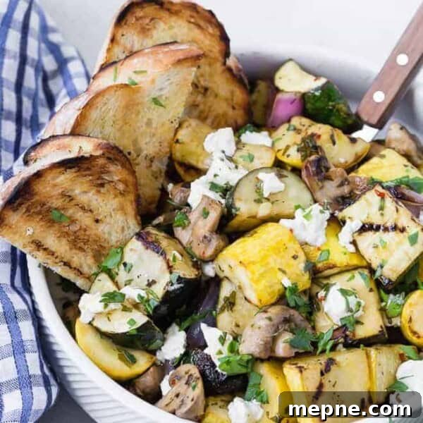 A white bowl filled with grilled zucchini, summer squash, mushrooms, and goat cheese. It is served with grilled bread pieces.