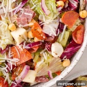 Close-up of a colorful salad featuring lettuce, cherry tomatoes, red onion, chickpeas, cheese cubes, sliced pepperoni, and shredded cheese in a speckled bowl.