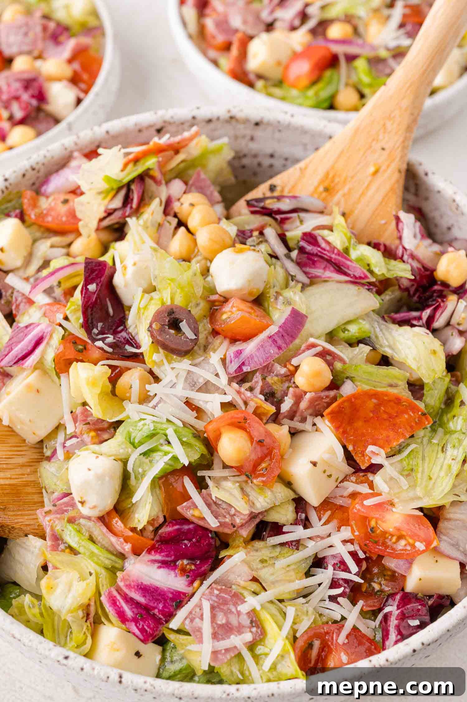 A mixed salad in a bowl featuring lettuce, tomatoes, cheese cubes, olives, chickpeas, and grated cheese, with a wooden spoon. Other bowls of salad are visible in the background.