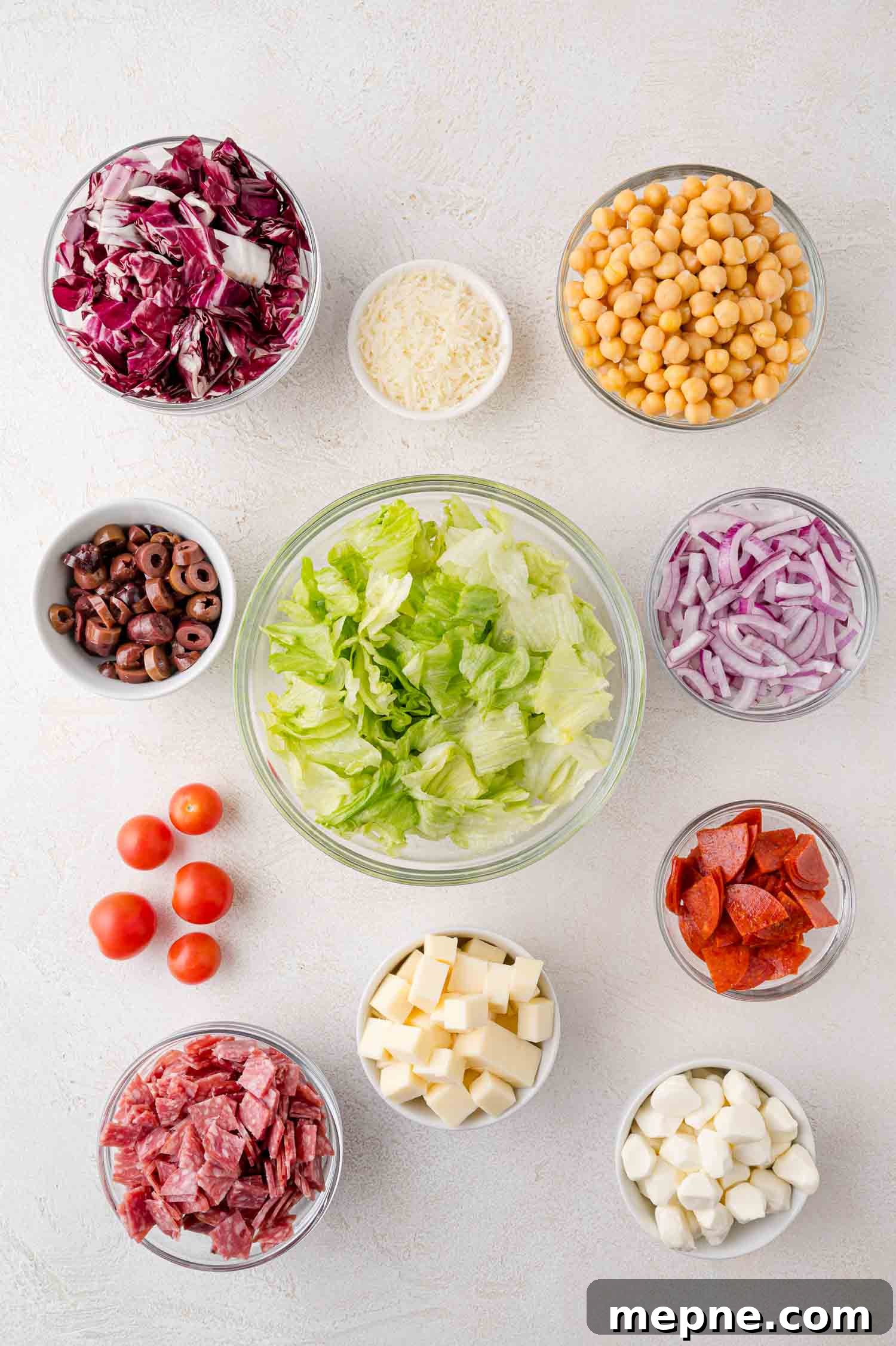 Bowls of salad ingredients including lettuce, chickpeas, radicchio, red onions, cheese, cherry tomatoes, olives, pepperoni, and salami, arranged on a white surface.