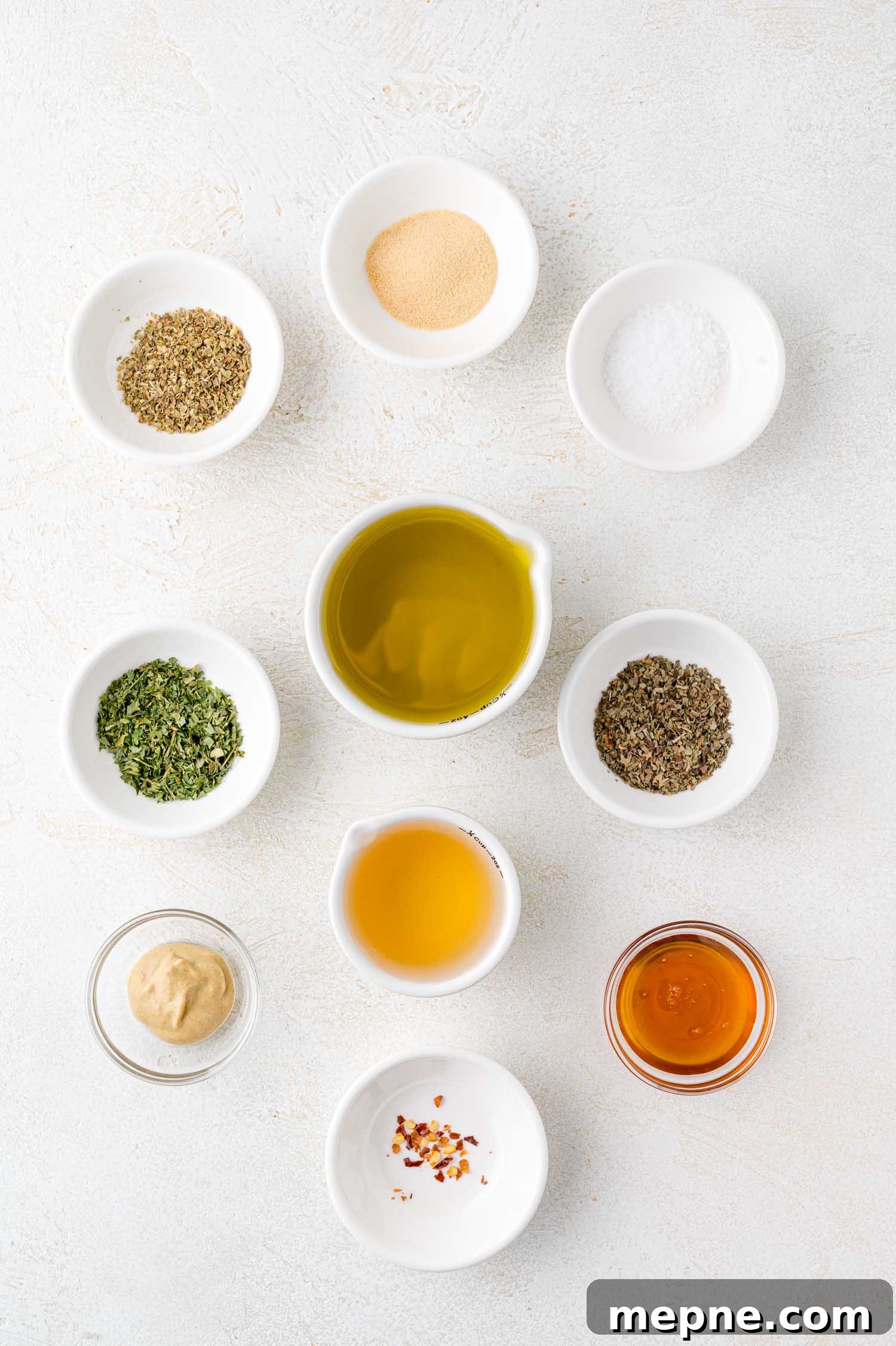 An assortment of small white bowls containing spices, herbs, oil, vinegar, mustard, and honey arranged on a white surface.
