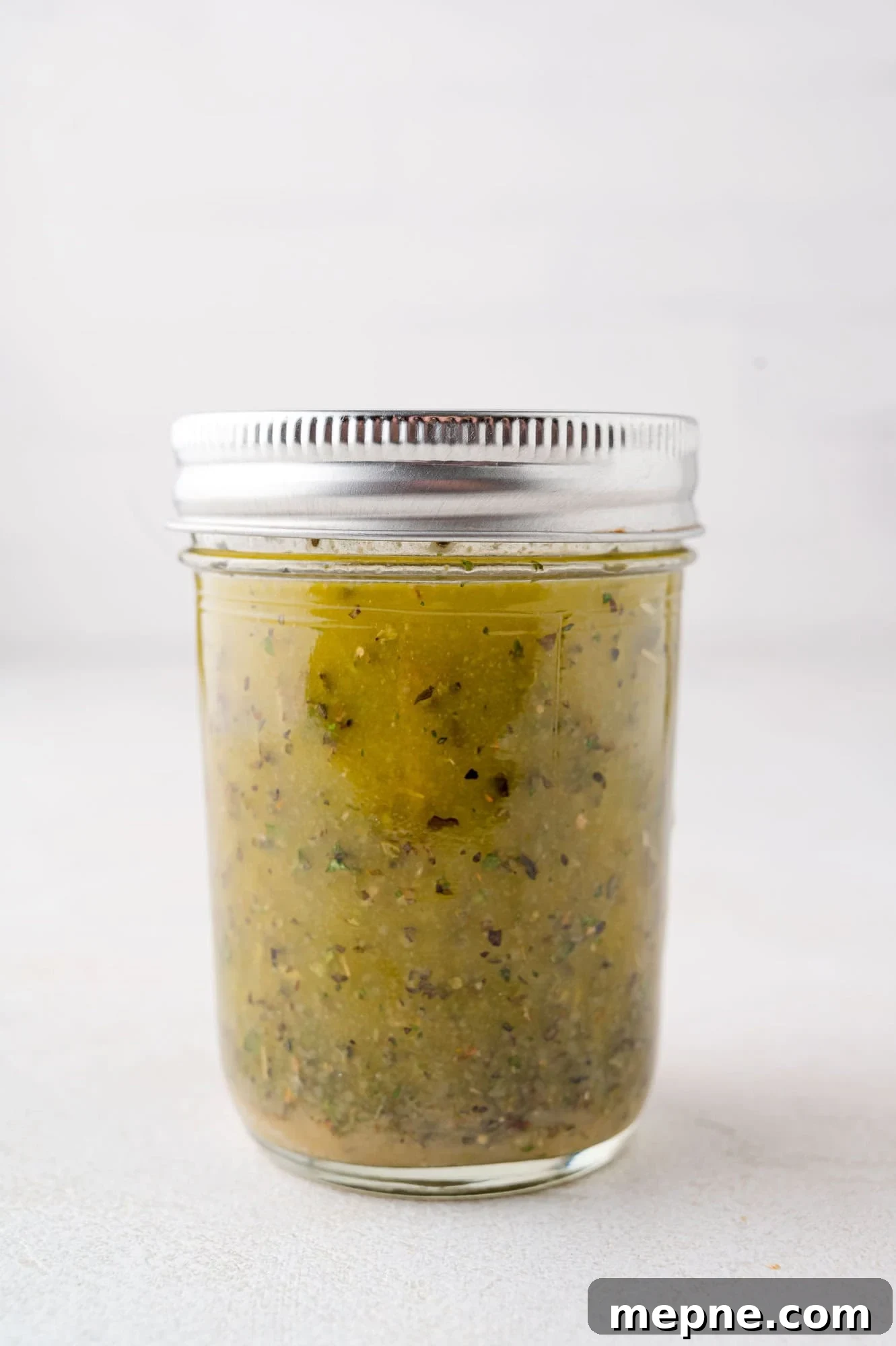 A clear, lidded jar filled with a greenish mixture that appears to be a sauce or condiment with visible herb and spice specks, standing on a light background.