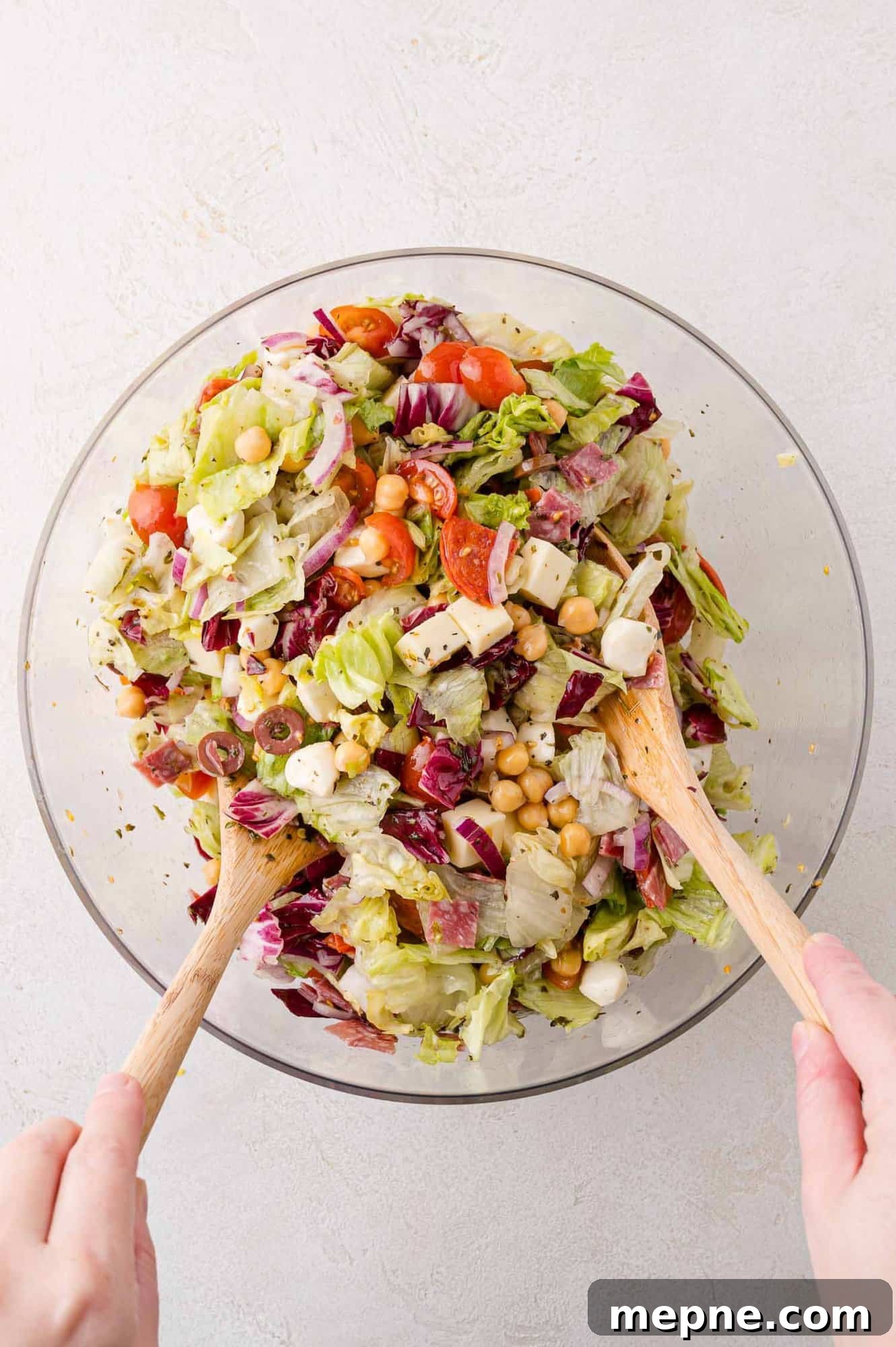 A person mixing a salad with wooden utensils in a glass bowl, containing lettuce, radicchio, chickpeas, mozzarella, olives, and cherry tomatoes.