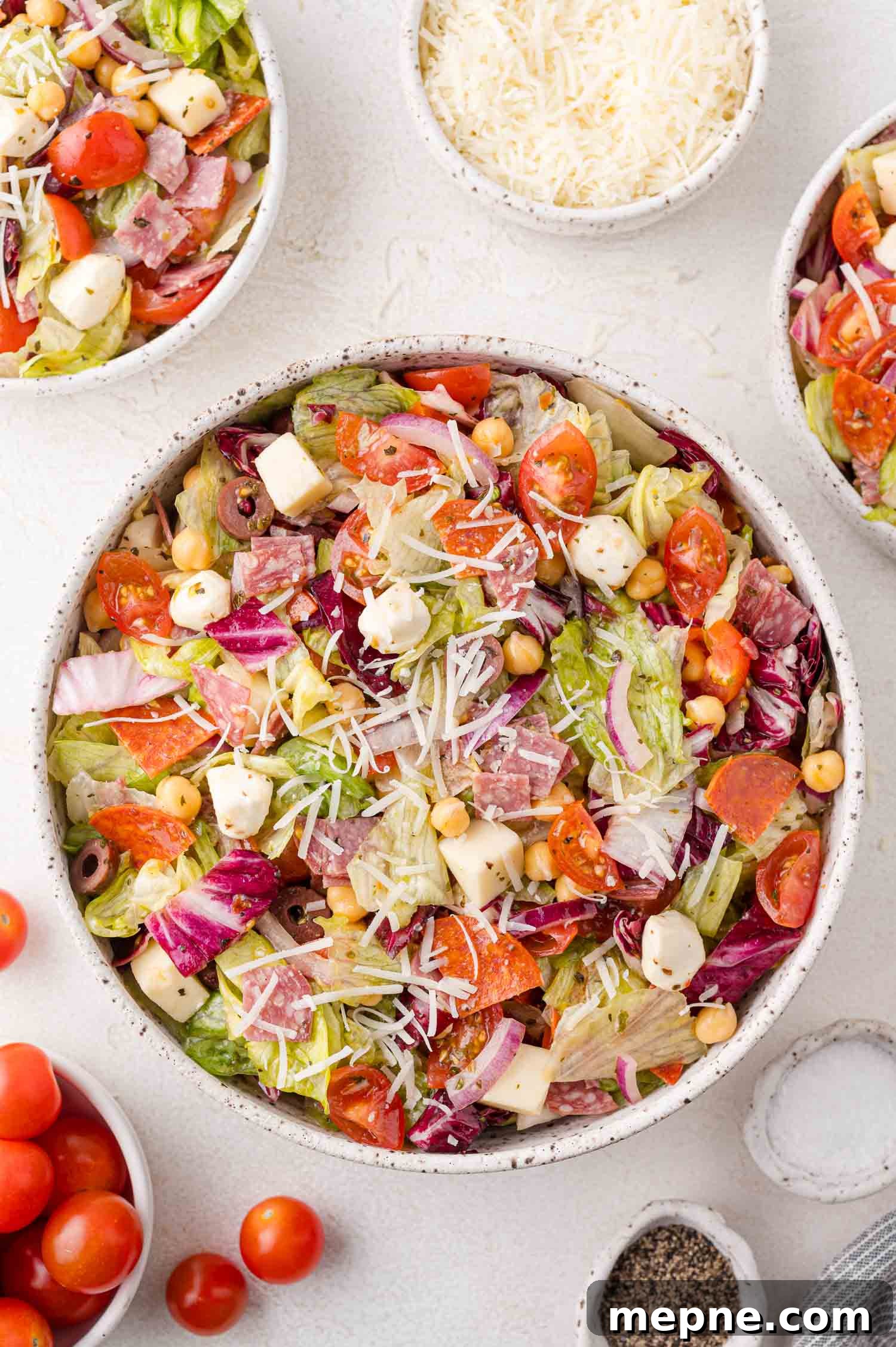A bowl of Italian chopped salad with lettuce, tomatoes, cheese, chickpeas, and cured meats, surrounded by small bowls of grated cheese, cherry tomatoes, and pepper.