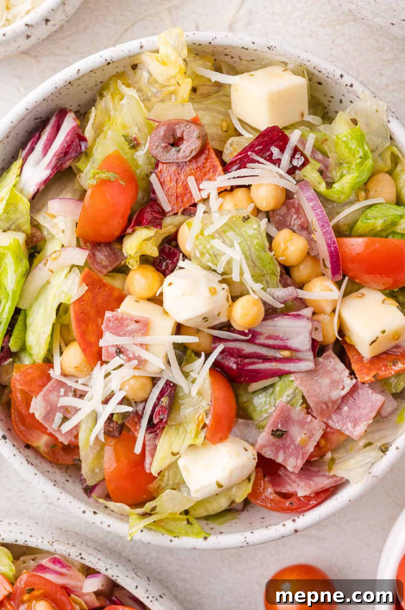 A bowl of salad containing lettuce, cherry tomatoes, red onion, chickpeas, mozzarella cubes, olives, salami, and shredded cheese.