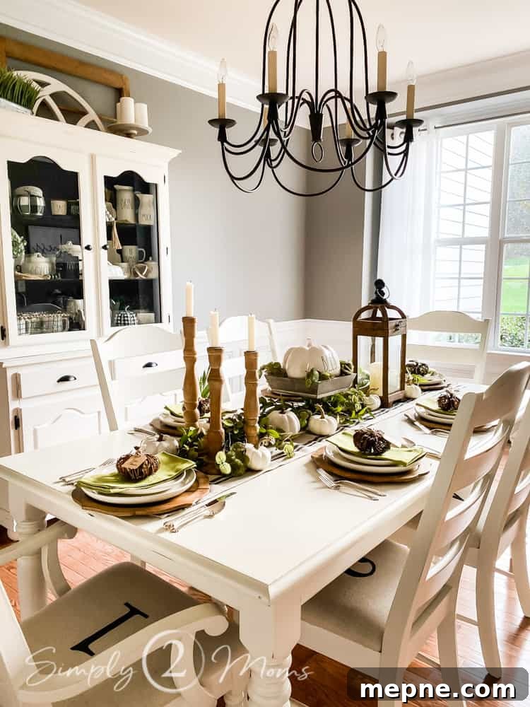 Painted rectangular dining room table with six painted ladder back chairs with fall tablescape. Farmhouse striped linen runner is on the center of the table running vertically. Centerpiece includes wood candlesticks with candles, large wood lantern with pillar candle, wooden square bowl on a pedestal with a large Rae Dunn pumpkin inside. Small white pumpkins and greenery fill in around all the larger items. Place settings include carved wood chargers, white dinner wear, folded green napkins topped with small wicker pumpkins. Painted china cabinet against the wall filled with Rae Dunn pottery and farmhouse baskets and buffalo check pumpkins. Wrought iron chandelier hangs over the table.