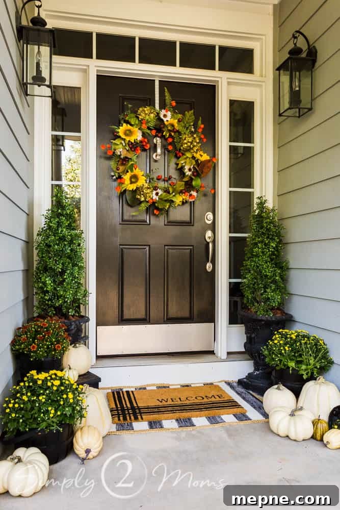 Painted black front door with sidelights and transom with brushed nickel kickplate, door knocker, and handle. A large fall floral wreath with sunflowers and greenery hangs on the door. Boxwood topiaries flank the door in large black urns with potted mums and white pumpkins on both sides. Coir doormat that reads welcome on top of a black and white striped rug.