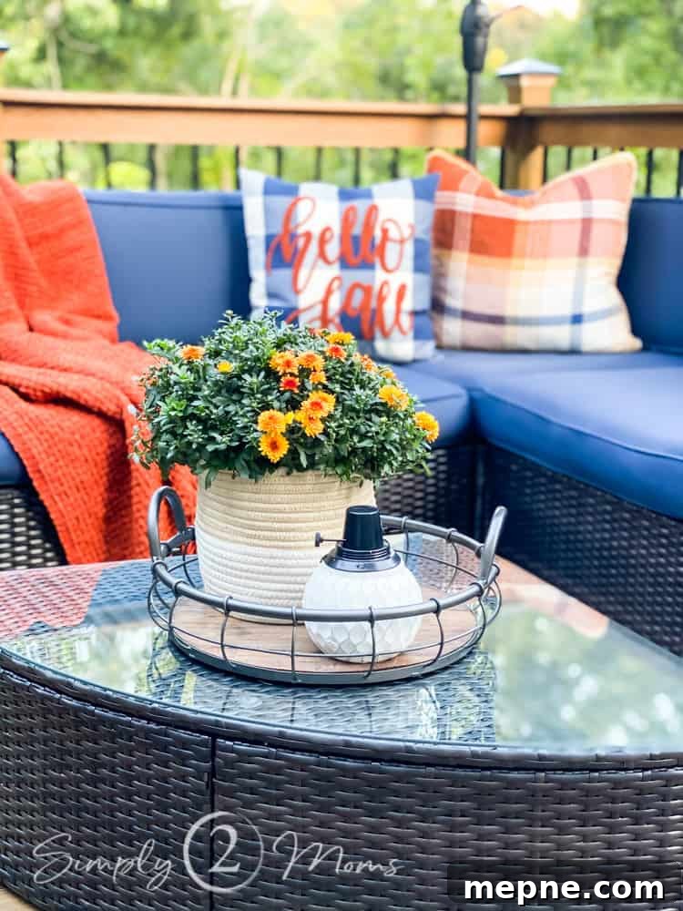 Outdoor sectional L-shaped wicker couch with blue box cushions and two plaid throw pillows in the corner. One pillow reads "hello fall" in script. Cozy throw blanket draped to left side. Wicker coffee table in front with a basket of mums and tray with pumpkin and lantern.