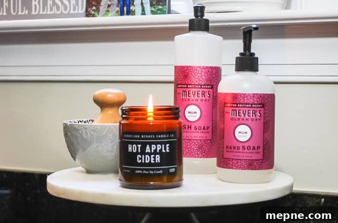 Pedestal cake stand with marble top holding Mrs. Meyer's hand soap and dish soap, a small bowl with a wood scrub brush and an amber glass jar with a candle inside.