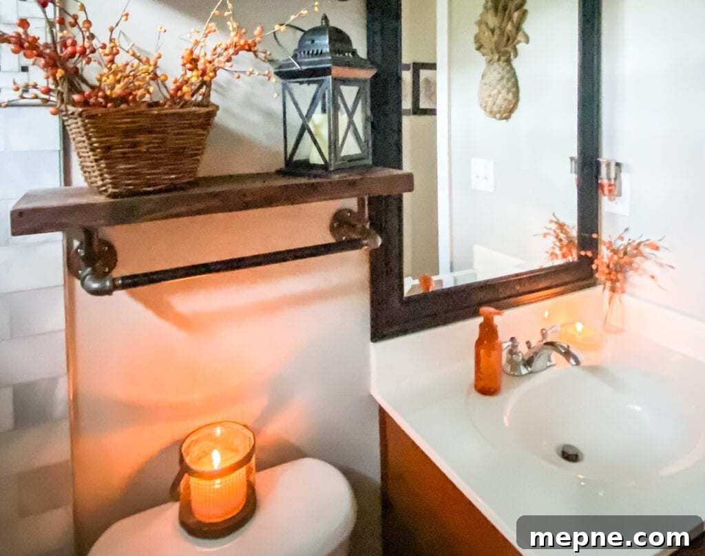 Bathroom with vanity holding fall stem, lit candle, and hand soap with pineapple hanging on the wall. Commode with lit candle and plumbing pipe shelf with fall berries and lantern.