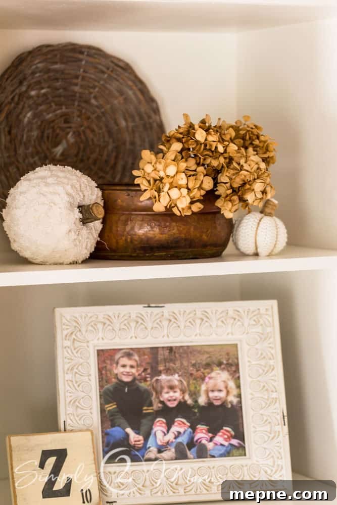 built in shelves with a copper container holding dried hydrangeas, and white sweater pumpkins. Wicker charger sits behind. Bottom shelf has a distressed white frame with picture of three young children wearing fall clothes with a large Scrabble letter with the letter Z on it.