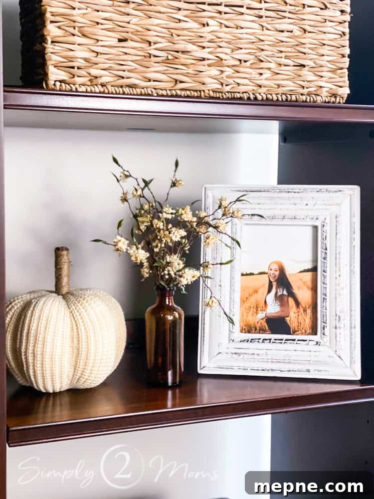Shelf of entertainment center with a white sweater pumpkin, small amber glass bottle holding a sprig of dried fall flowers, and a distressed farmhouse style picture frame holding a photo of a teenage girl standing in a field of dried grass