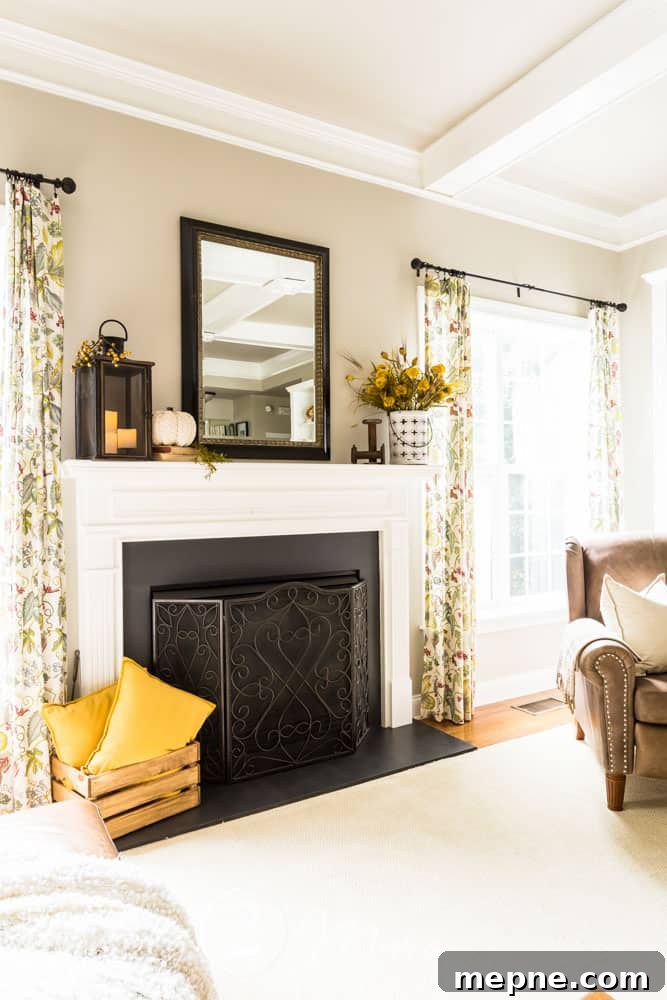 Fireplace with mantel made from moldings and black chalk painted marble surround with scrollwork iron fireplace screen. Mantel has a large lantern with three candles on one side, a woven pumpkin is next to the lantern on top of two vintage books. Large-scale black framed mirror is hanging centered above the fireplace. White lantern bucket filled with a fall floral arrangement is on opposite side of mantle with vintage textile spools placed next to it. Wood crate filled with two pillows sits on the hearth.