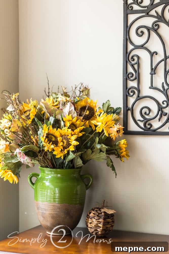Large urn-style pottery vase with clay bottom and glazed top filled with a bouquet of sunflowers, fall greenery, and twigs. A woven pumpkin sits next to the vase. An iron scrollwork gate hangs on the wall.