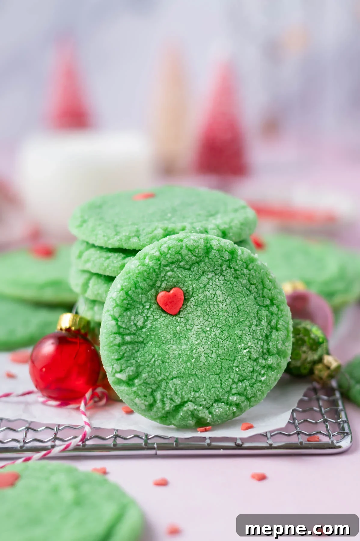 Soft Chewy Grinch Sugar Cookies for Christmas 2 grinch cookie leaning up against a stack of cookies with milk and Christmas trees in the background