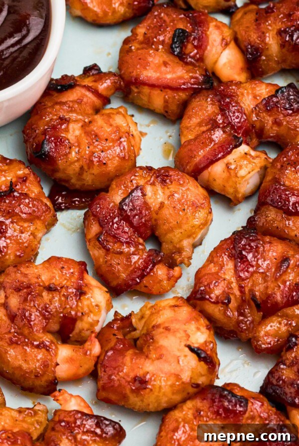 Crispy bacon wrapped around shrimp, coated with BBQ sauce mixture, on a light green plate with lemon slices and serving tongs on the table.