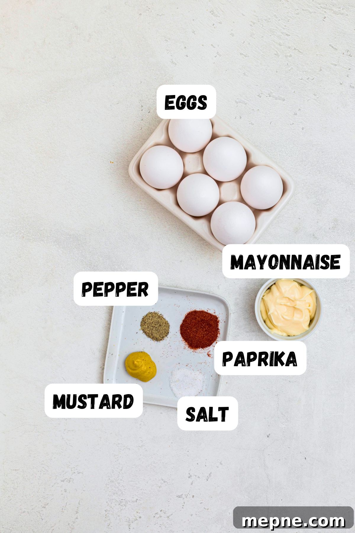 Eggs, mayo, mustard, salt, pepper, and paprika, measured out and labeled on a white marble table.