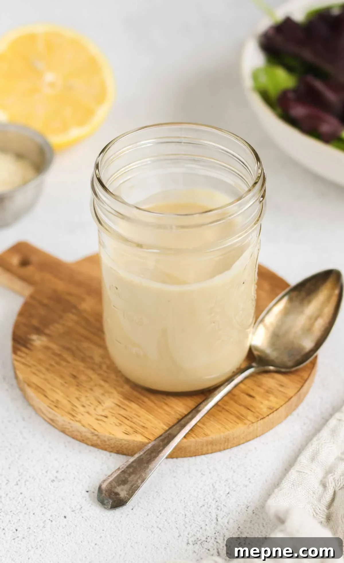 Creamy Maple Tahini Dressing for Salads and Grain Bowls 2 Maple tahini dressing in a mason jar with a spoon on the side of the jar and a lemon and bowl of mixed greens in the background.