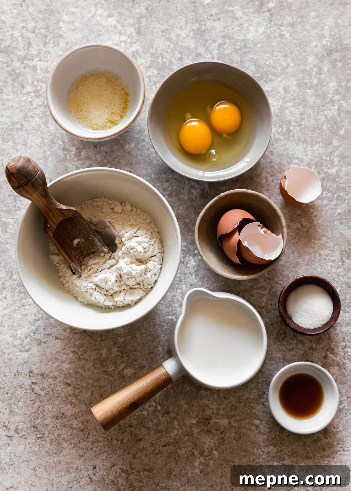 An overhead image of a white bowl of flour with a wood scoop, a cream bowl of egg shells, a white cup of milk, a wood bowl of salt, a beige bowl of eggs, and a white bowl of melted butter on a beige counter.