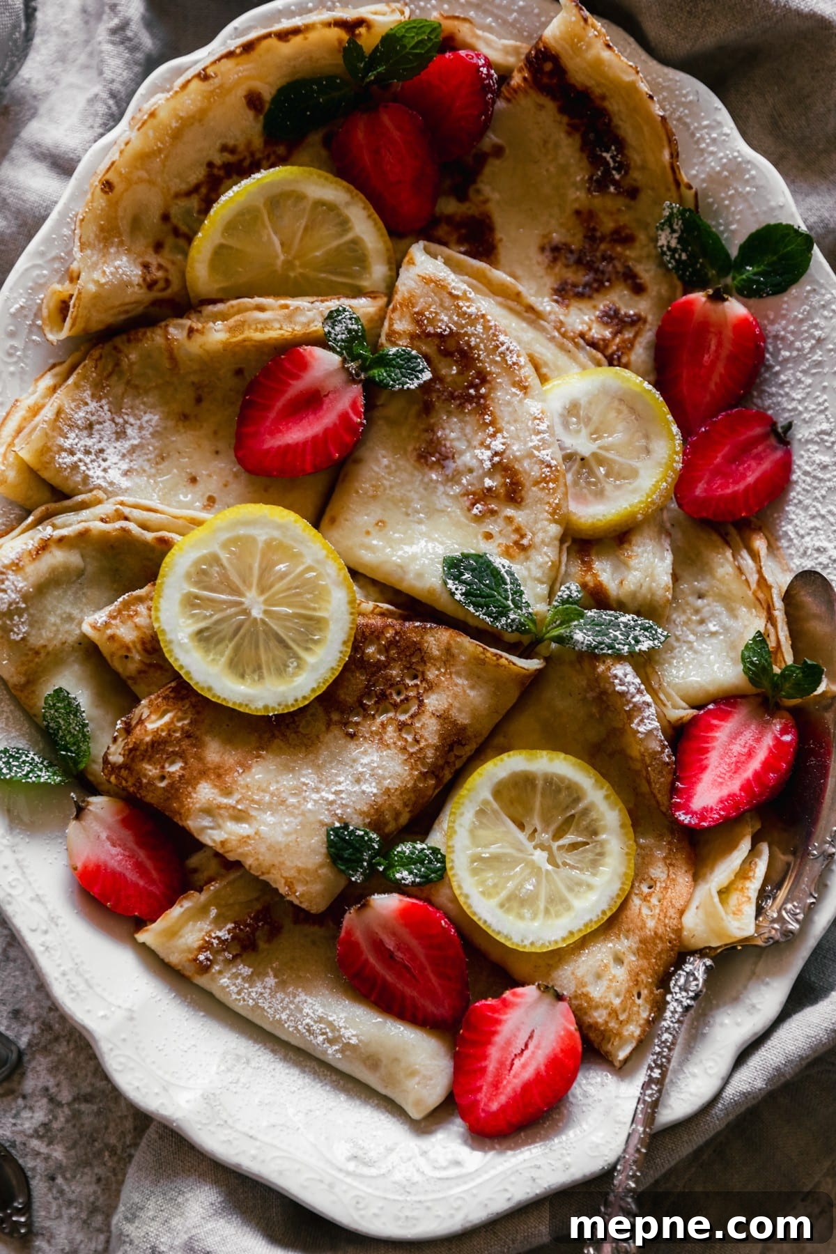 A very closeup image of Norwegian pancakes with strawberries and lemon slices on a white oval plate with a vintage serving spoon in the right lower corner. The background is a beige linen and tan speckled table.