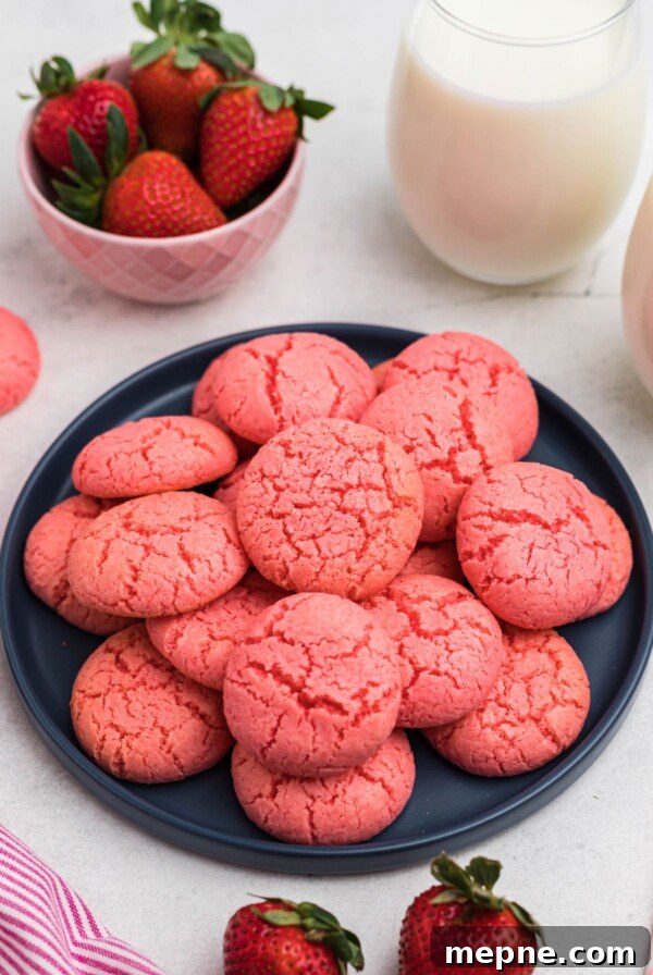 3 Ingredient Sugar Cookies Recipe 10 Pink strawberry cake mix cookies stacked on a blue plate in front of glasses of milk and bowls of strawberries.