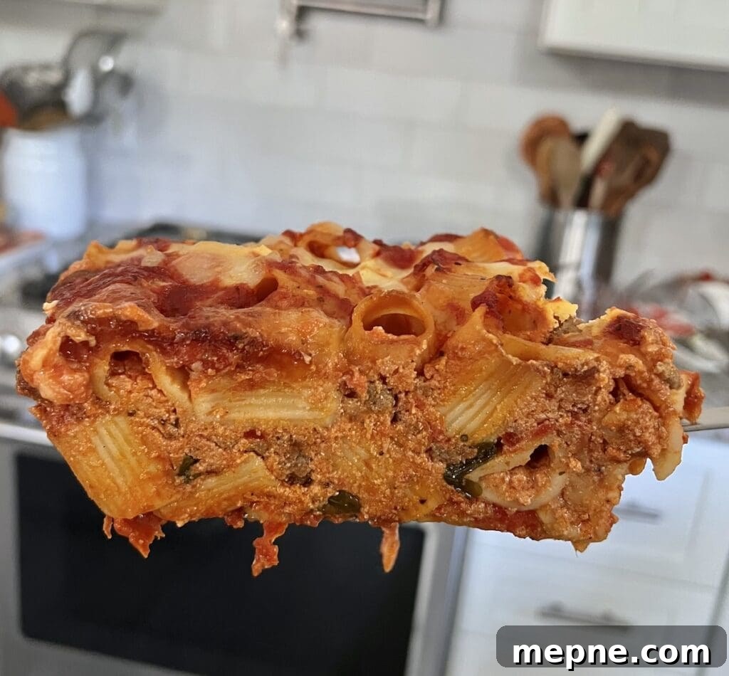 Side view of a large slice of pasta al forno on a spatula with kitchen in the background.