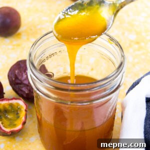 A spoon drizzles passion fruit sauce into a mason jar sitting on a yellow surface surrounded by halved purple passion fruits