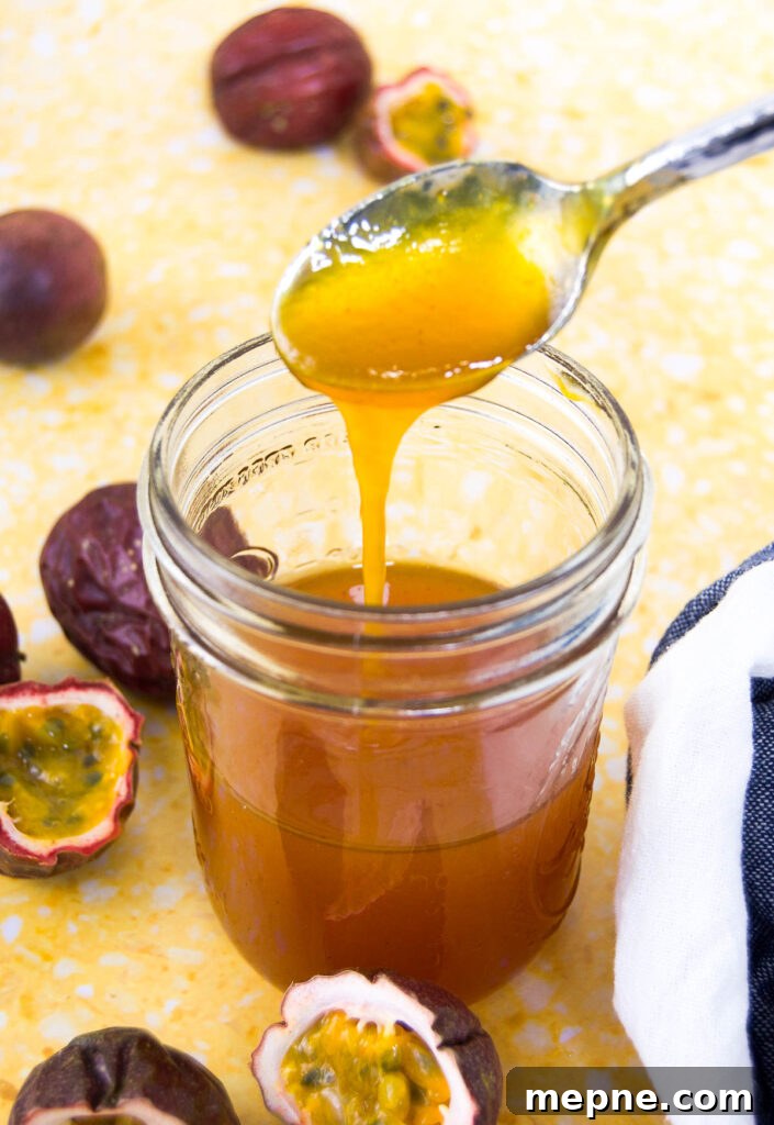 Homemade Passion Fruit Sauce Recipe for Desserts 5 A spoon drizzles passion fruit sauce into a mason jar sitting on a yellow surface surrounded by halved purple passion fruits