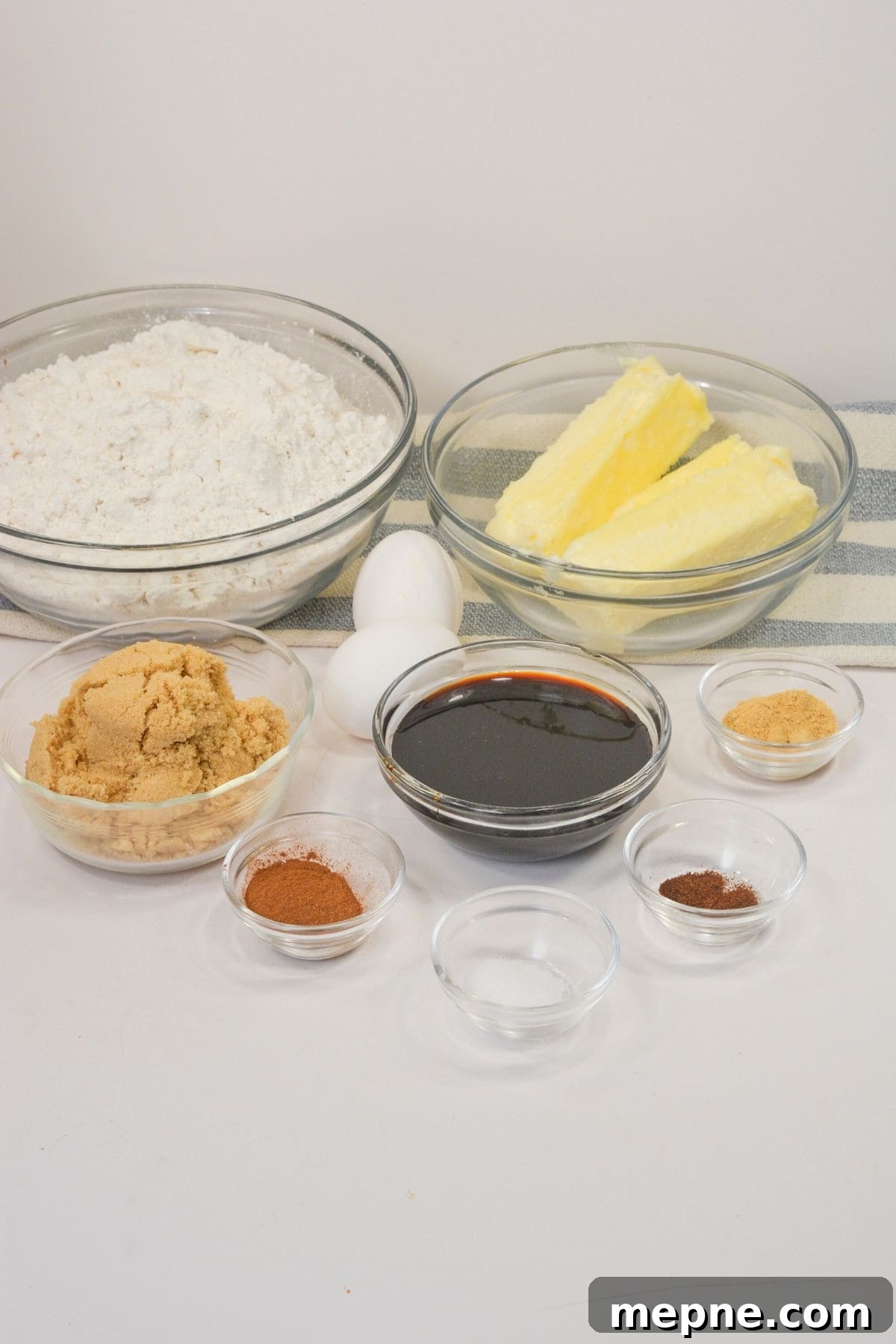 Homemade Soft Chewy Ginger Molasses Cookies Recipe 4 A variety of baking ingredients for Ginger Molasses Cookies are arranged in clear bowls, including flour, brown sugar, butter, eggs, molasses, and several spices on a white surface.
