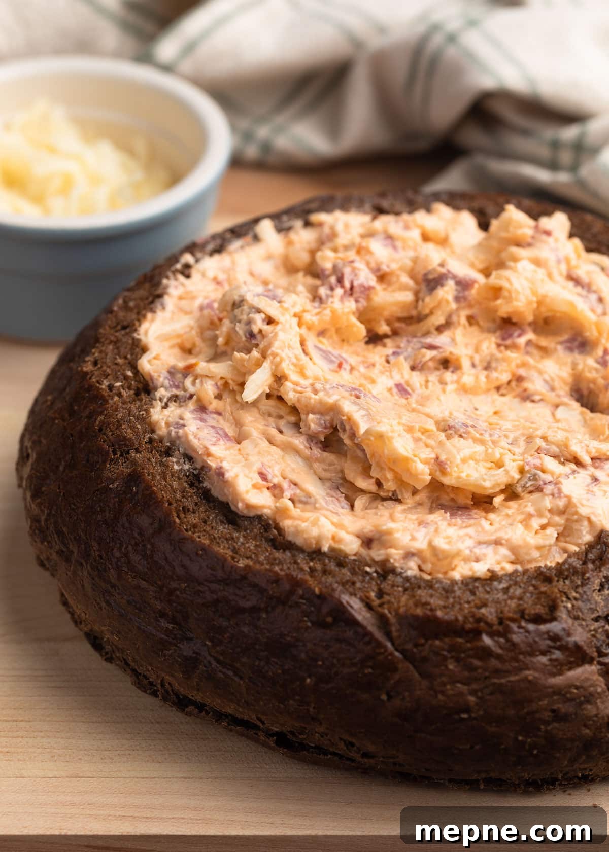 unbaked reuben dip in a pumpernickel bread bowl on a light wood cutting board with a bowl of shredded swiss cheese on the side