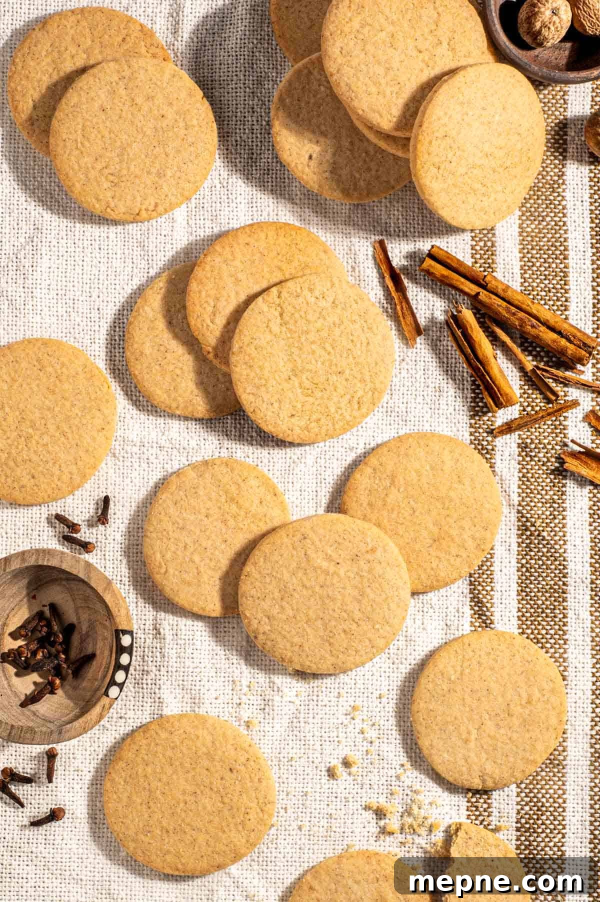 Old Fashioned South African Spice Biscuits (Soetkoekies) Recipe 2 Outydse soetkoekies scattered across a textured tea towel, surrounded by nutmeg, cloves and cinnamon sticks.