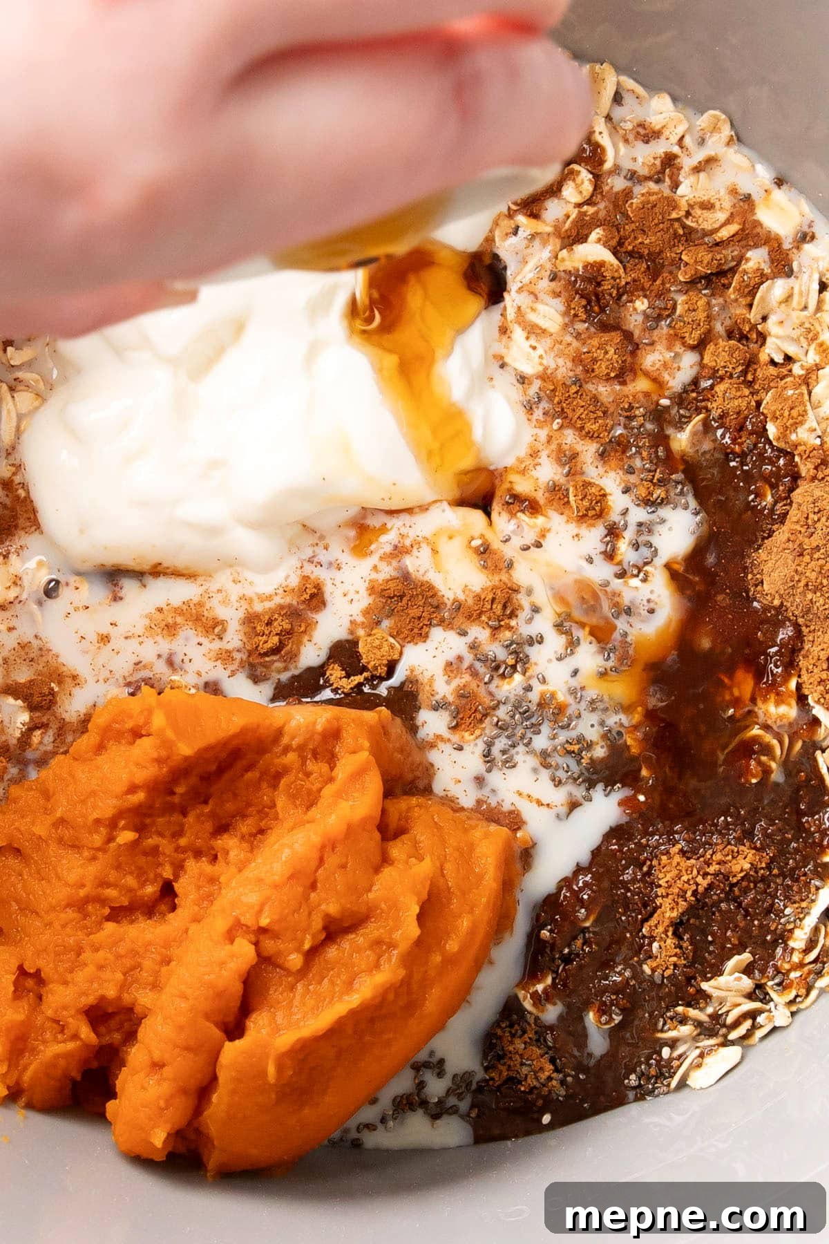 Pumpkin pie overnight oats ingredients in a mixing bowl including chia seeds, rolled oats, cinnamon, pumpkin pie spice, pumpkin puree and someone adding maple syrup