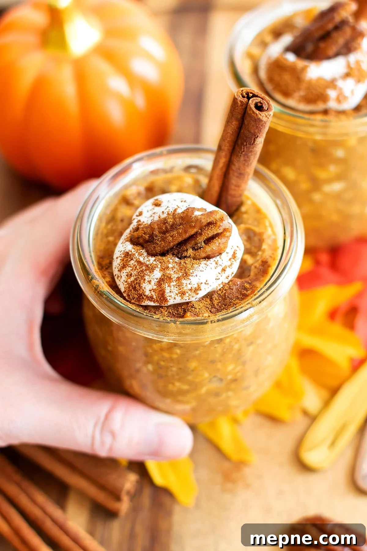 Someone holding a jar of pumpkin chia overnight oats topped with a cinnamon stick and whipped cream with a pecan