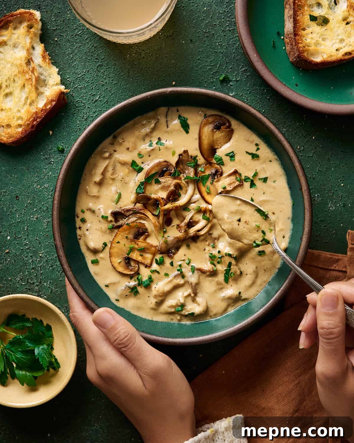 woman's hands spooning creamy vegan mushroom soup from a green bowl on a green table.
