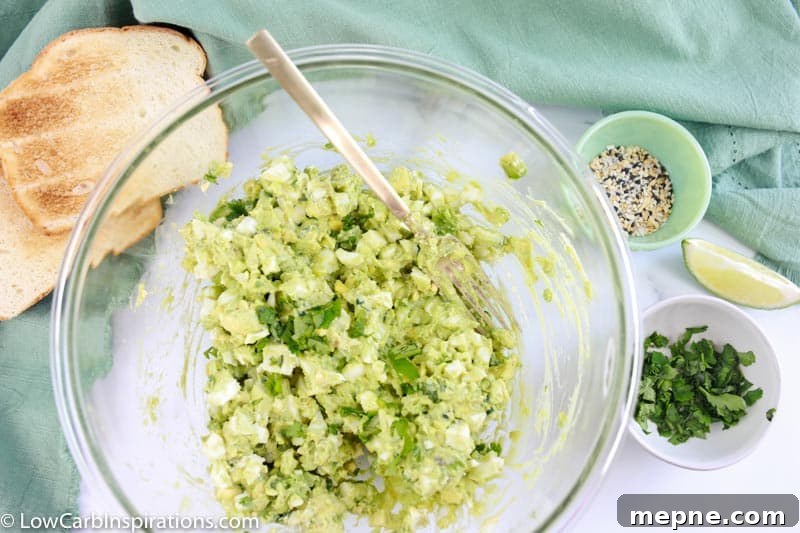 mixed avocado egg salad recipe in a clear bowl with extra seasonings and low carb bread on the side
