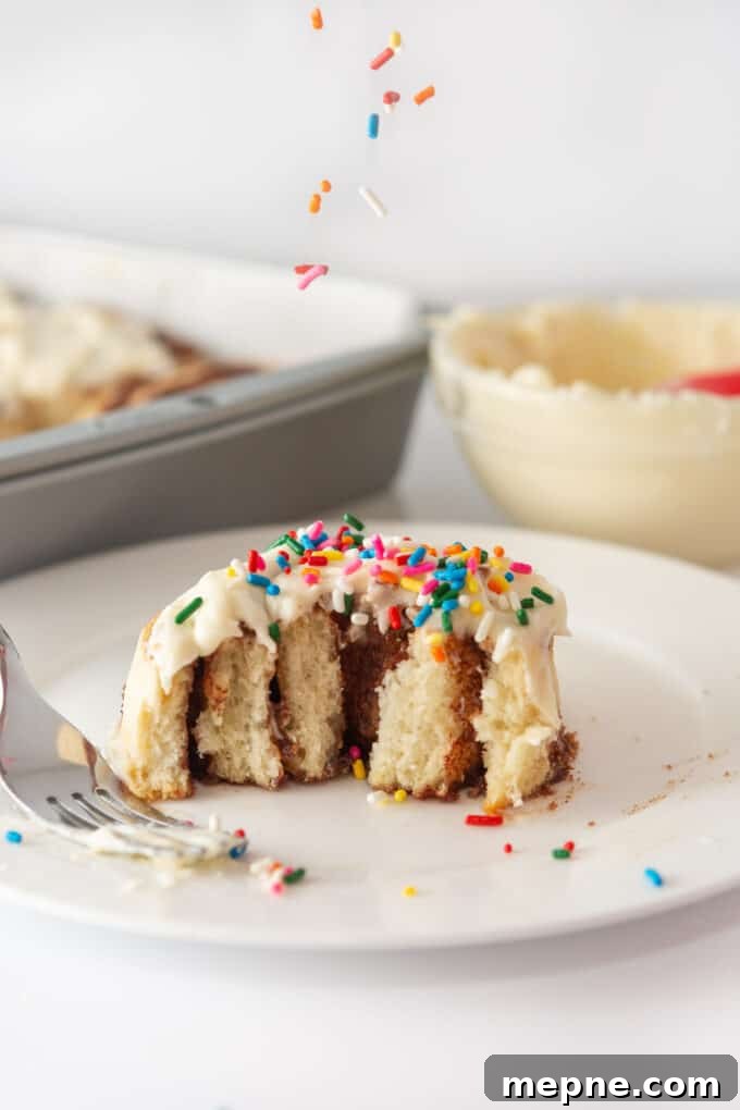 Cream Cheese Frosting Recipe for Cinnamon Rolls 5 A single cinnamon roll on a white plate, pulled apart to reveal its layers, topped with cream cheese frosting and rainbow sprinkles