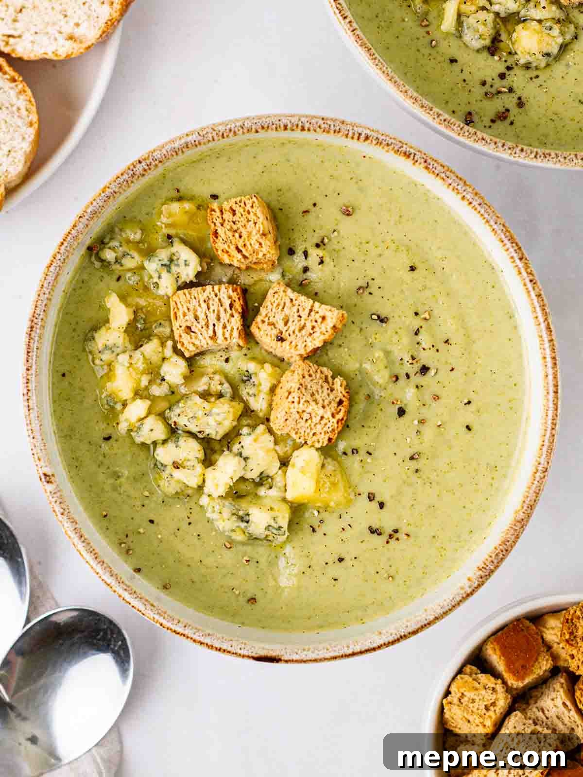 Creamy Broccoli and Stilton Soup Recipe 2 A bowl of Broccoli and Stilton Soup topped with cheese and croutons on a table with soup, bread and spoons.