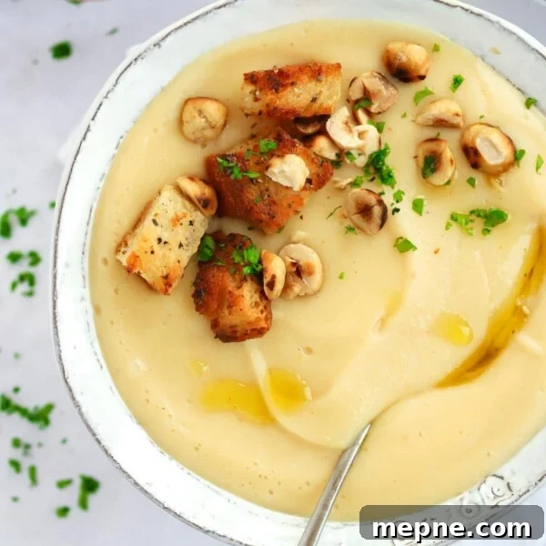 Creamy Broccoli and Stilton Soup Recipe 9 A bowl filled with creamy parsnip soup, with a spoon in and garlic croutons and halzenuts sprinkled on top. The bowl is on top of a white tablecloth.
