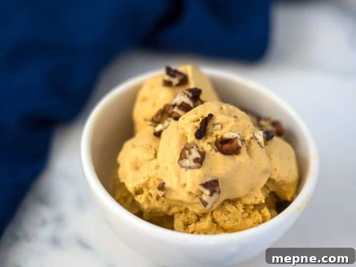 25 Low Carb Ice Cream Recipes for Creamy Keto Desserts 16 A white bowl filled with scoops of pumpkin cheesecake ice cream topped with chopped pecans, placed on a marble surface with a blue napkin in the background.