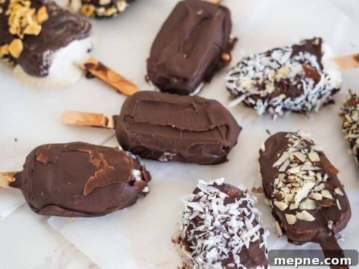 25 Low Carb Ice Cream Recipes for Creamy Keto Desserts 21 Assorted chocolate-covered ice cream bars on sticks, topped with various toppings such as nuts, coconut flakes, and chocolate drizzle, placed on a white paper-lined surface.