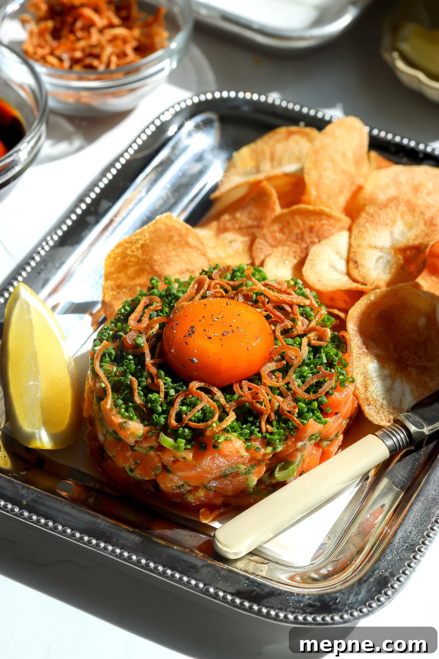 Spicy Salmon Tartare with Soy-Cured Egg and Crispy Chips Recipe 2 A beautifully plated spicy salmon tartare on a silver platter, garnished with a glossy soy-cured egg yolk, fresh chives, and crispy fried shallots, served with homemade potato chips.