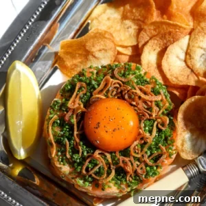 An overhead view of a spicy salmon tartar appetizer on a silver tray, showing the cured egg yolk and crispy toppings with lemon wedges.