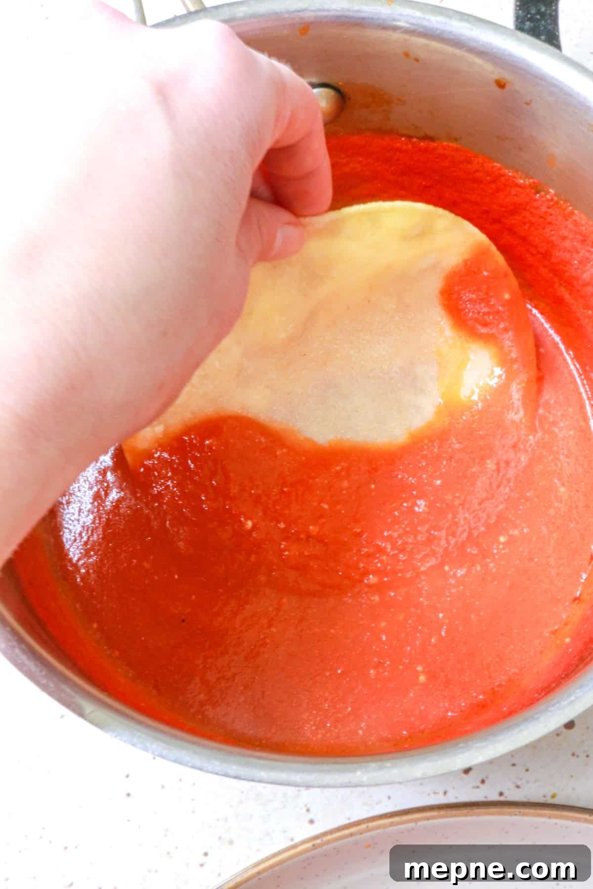 a hand dipping a corn tortilla in tomato sauce