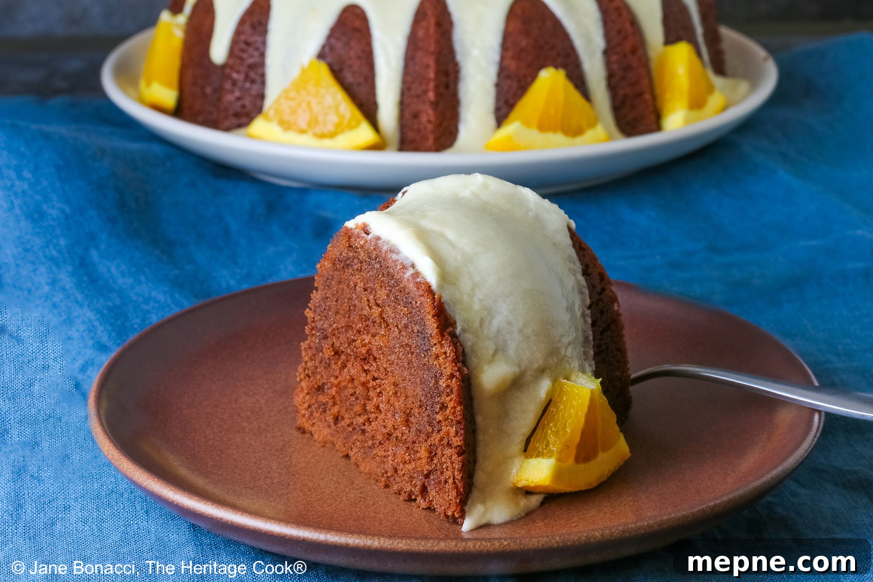 Whole chocolate/orange bundt cake with cascading orange flavored glaze and fresh orange slices around the exterior on a white plate, sometimes on its own, sometimes with a slice cut out and on a plate in front, on a blue cloth with a dark gray background © 2026 Jane Bonacci, The Heritage Cook.