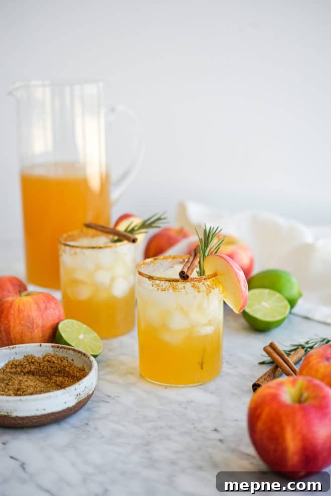 Spiced Apple Cider Margarita Recipe 4 two apple cider margaritas next to a pitcher of margaritas next to apples, limes, brown sugar, cinnamon sticks, and rosemary